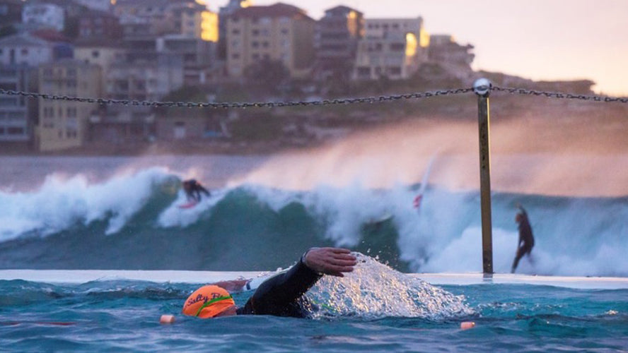 swimmer in outdoor pool with surfers in the sea behind 