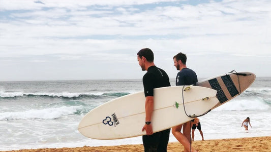 Two surfers walking towards to sea wearing wetsuits and holding surfboards 