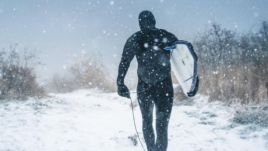 A surfer wearing neoprene accessories and carrying a surfboard in the snow