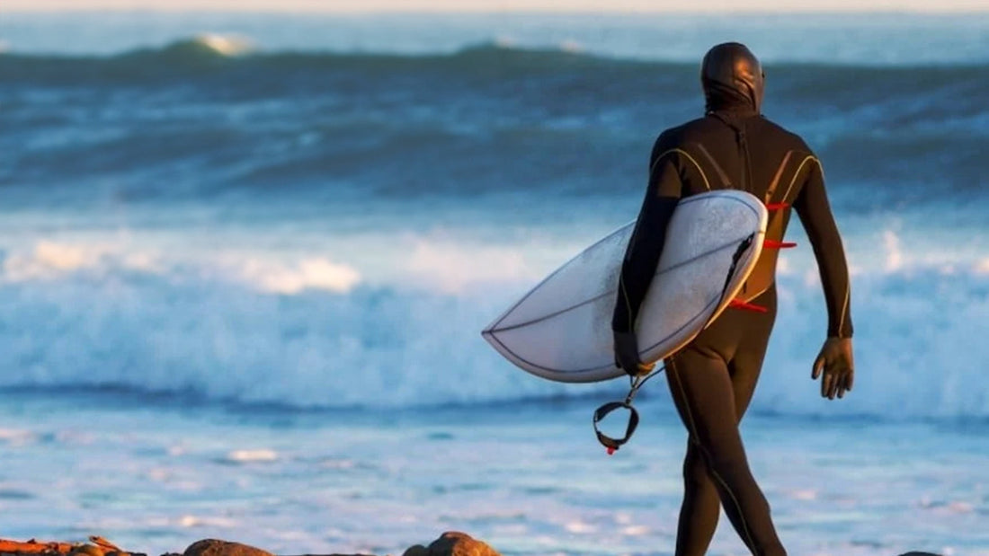 A surfer in hooded winter wetsuit carrying a surfboard